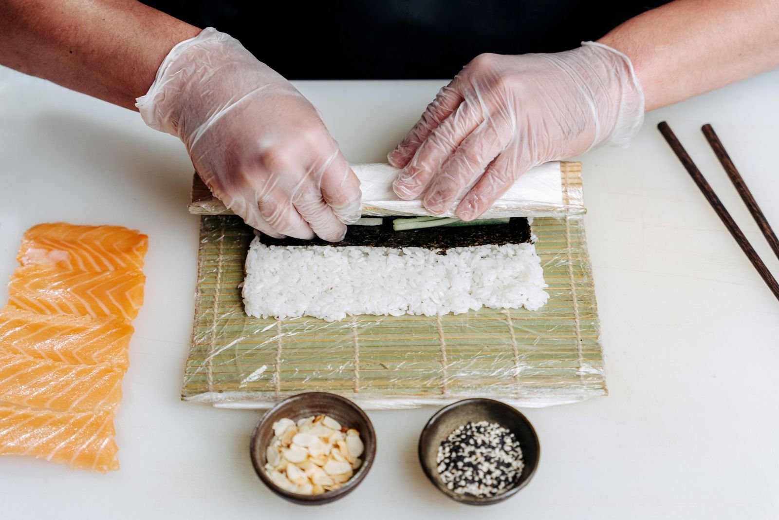 Gloved chef rolling sushi with salmon, rice, nori and sesame ingredients